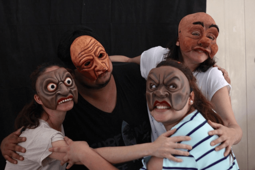 Four people wearing commedia dell'arte character masks have their arms around each other in front of a black curtain.